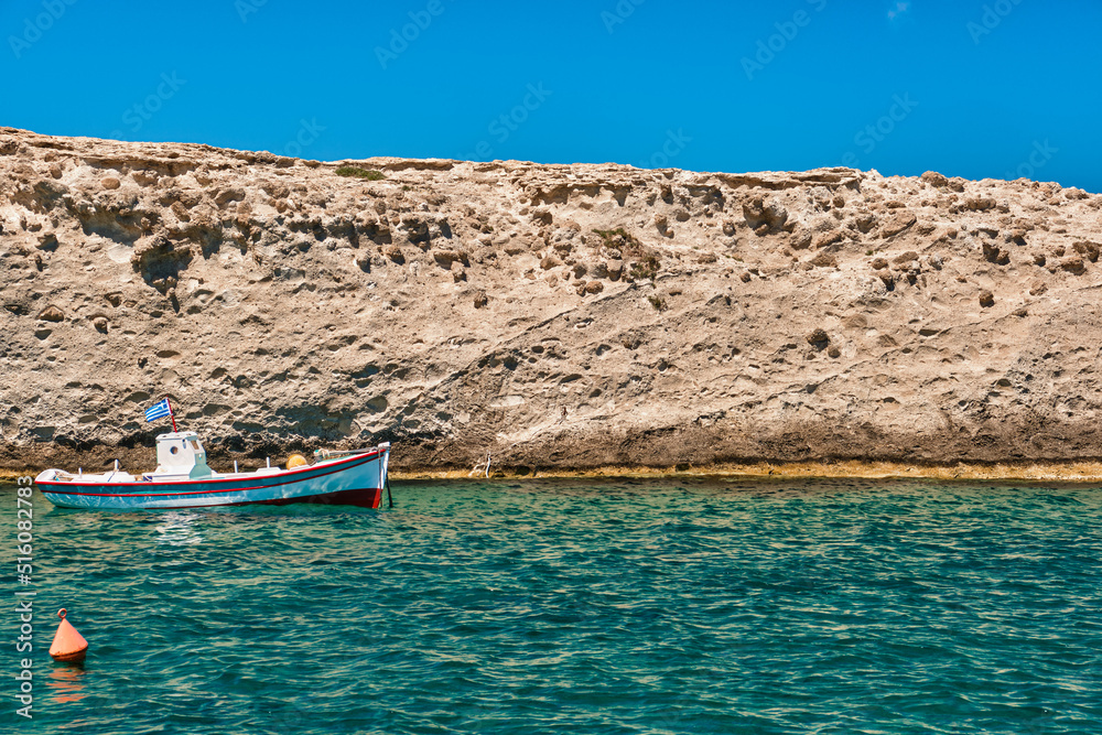 Fototapeta premium Anchored small fishing boat under Greek flag, azure sea waters, clear blue sky, rocky hill, red fishing buoy floating. Frontal shot. Milos, Greece.