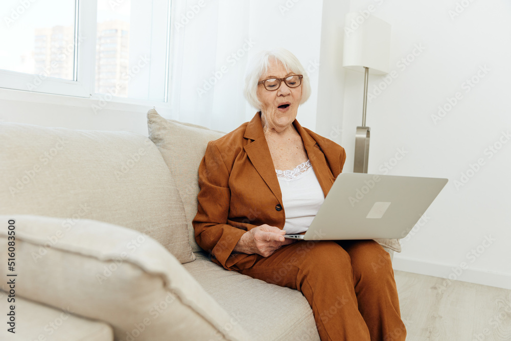 a pleasant, attractive elderly woman in a brown suit is sitting on a beige sofa working on a laptop remotely, being in a comfortable environment a pleasant, attractive elderly woman in a brown suit is sitting on a beige sofa working on a laptop remotely, being in a comfortable environment