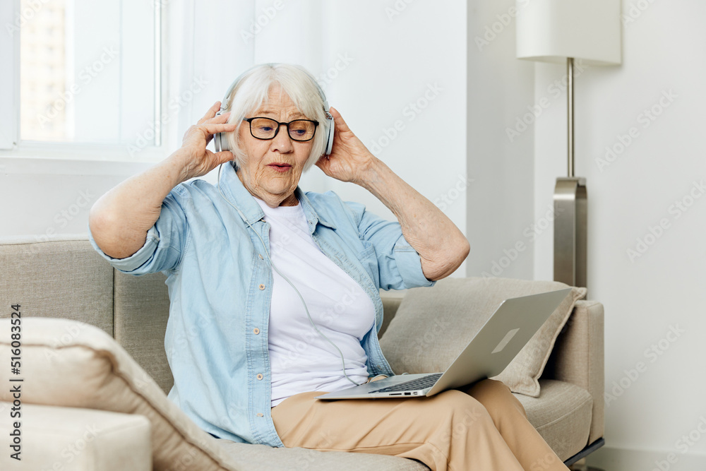 a pleasant joyful elderly lady uses modern technologies holding a laptop on her lap and holding headphones with her hands a pleasant joyful elderly lady uses modern technologies holding a laptop on her lap and holding headphones with her hands