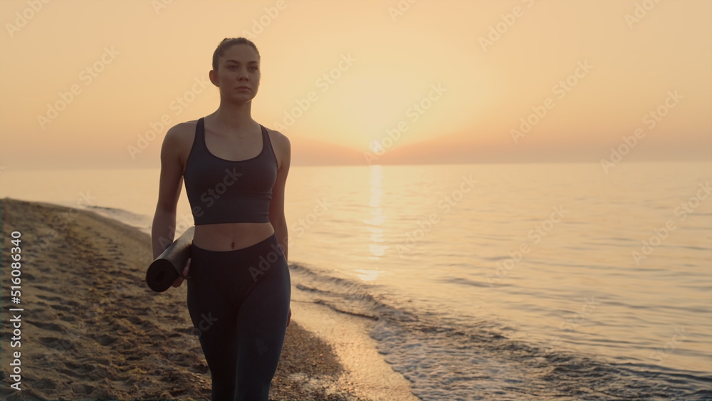 Athletic woman walking beach holding sport mat at sunset. Girl going exercising.