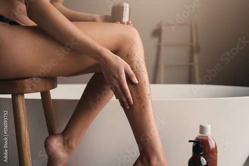 Cropped image of female hand applying peeling coffee scrub, massaging legs in bathroom photo