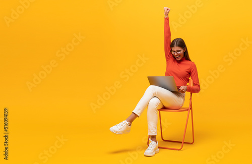 Excited young woman sitting on chair with laptop, shouting yes as winner, isolated on yellow