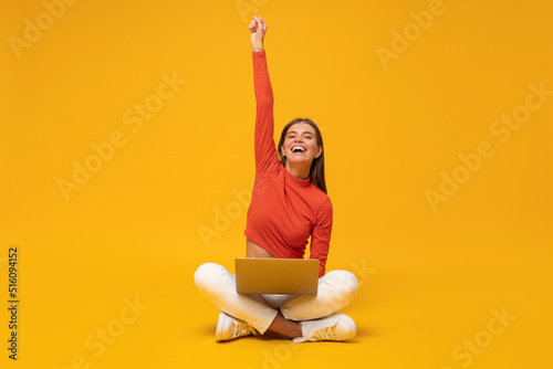 Excited female student sitting on floor with laptop, raising hand as a winner, isolated on yellow photo