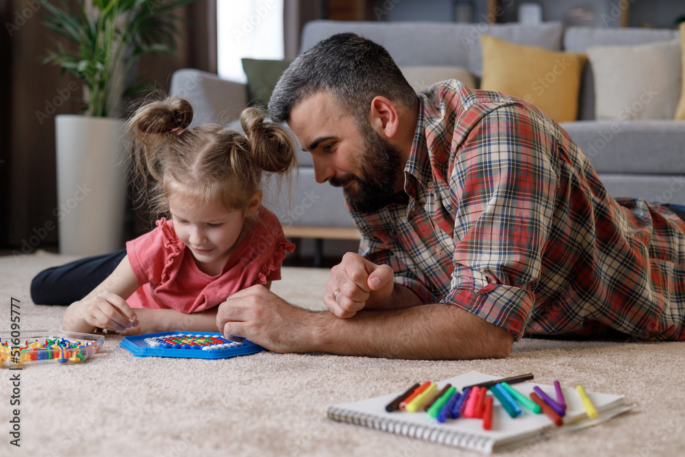 Young handsome father and cute daughter playing together. Dad and child making a puzzle with mosaic pegboard lying on the carpet at home. Educational toys for children. Home leisure. Father's day Young handsome father and cute daughter playing together. Dad and child making a puzzle with mosaic pegboard lying on the carpet at home. Educational toys for children. Home leisure. Father's day