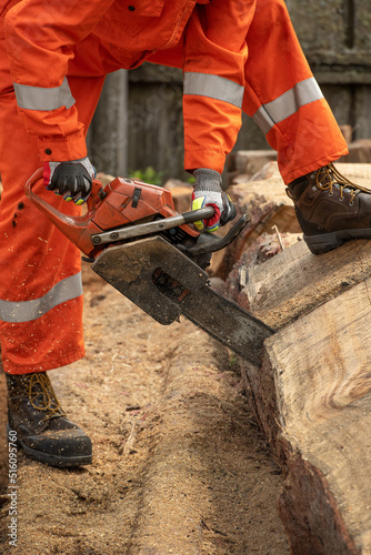 Close up of chain saw used cut log. Body of workmen in orange coveralls.