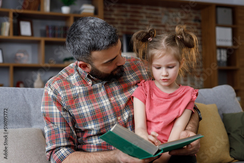 Young father and daughter read intersting book at home. Loving parent bearded man teaches his preschool child to read. Daddy reading fairy tale story. Parenthood, love and happy family concept photo