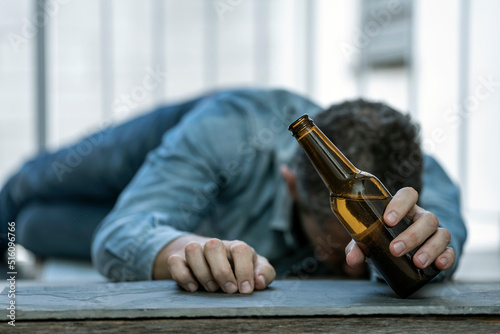 DRUNK MAN LYING ON THE FLOOR IN THE STREET ASLEEP WITH A BOTTLE OF BEER IN HIS HAND. ALCOHOL CONSUMPTION ADDICTION. ALCOHOLISM CONCEPT. FOCUS SELECTED.