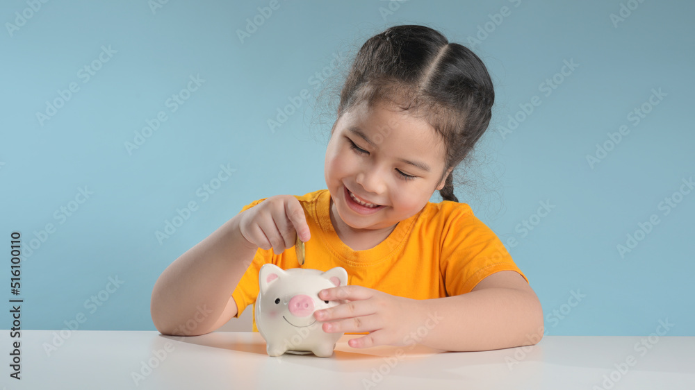Asian cute girl saving money putting coins into piggy bank on table at home, learning about saving, Kid save money for future education. Money, finances, insurance, and people concept Asian cute girl saving money putting coins into piggy bank on table at home, learning about saving, Kid save money for future education. Money, finances, insurance, and people concept