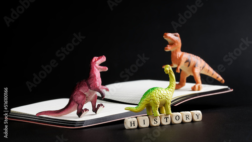 Three toy dinosaurs stand on an open notebook next to the inscription history. The concept of studying history, the Mesozoic era and ancient reptiles. Black background. Copy space. photo