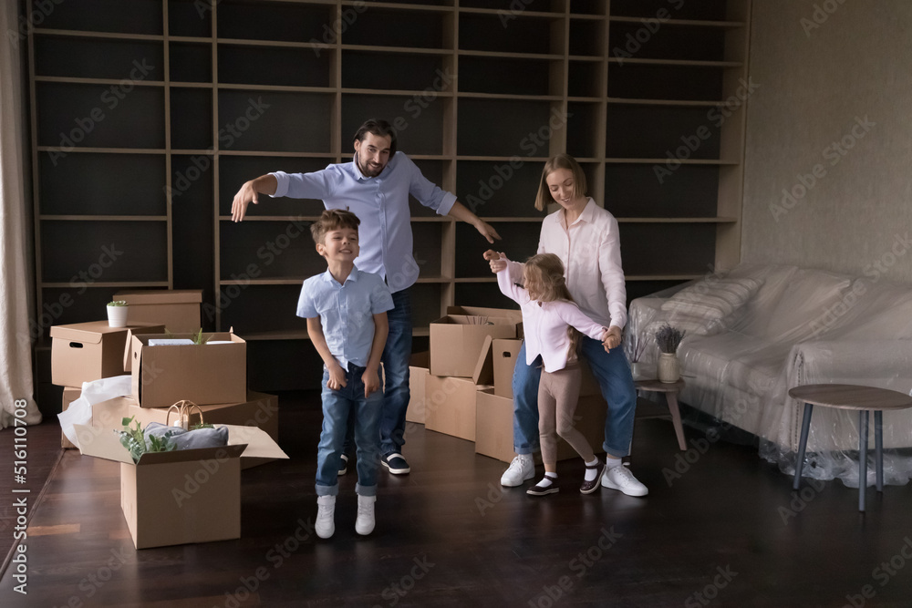 Happy active parents teaching two energetic kids to dance at new home, enjoying family party at moving paper boxes, smiling, laughing, having fun, celebrating, house buying, apartment rent, renovation