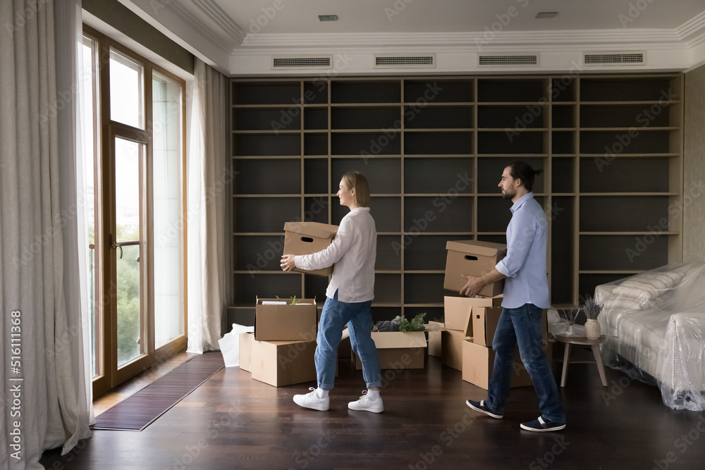 Young millennial married couple carrying boxes into empty living room, moving into new home after renovation, renting or buying apartment. Relocation, real estate concept. Full length shot