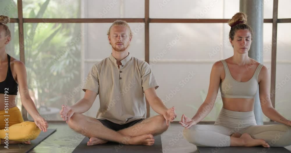 Pan-shot of group of five young, healthy, caucasian man and woman closed eyes sitting on exercise mat, practicing yoga meditation in class together at fitness studio. Health and Wellness Concept.