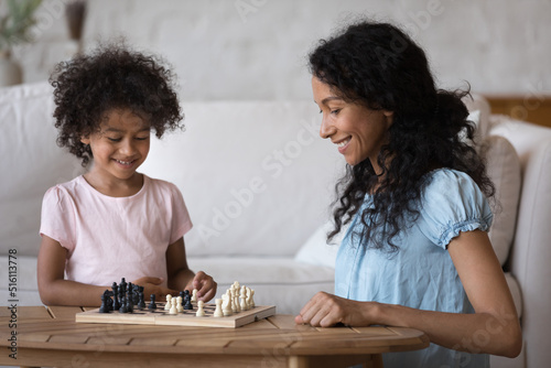 In living room African mother engaged in board game with cute little daughter, family enjoy chess hobby activity, spend leisure at home. Education, development, teach creative logical thinking concept