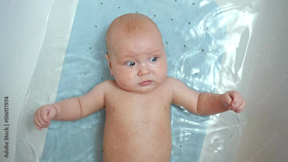 Baby girl lies in bathtub with water. Child looks around with little