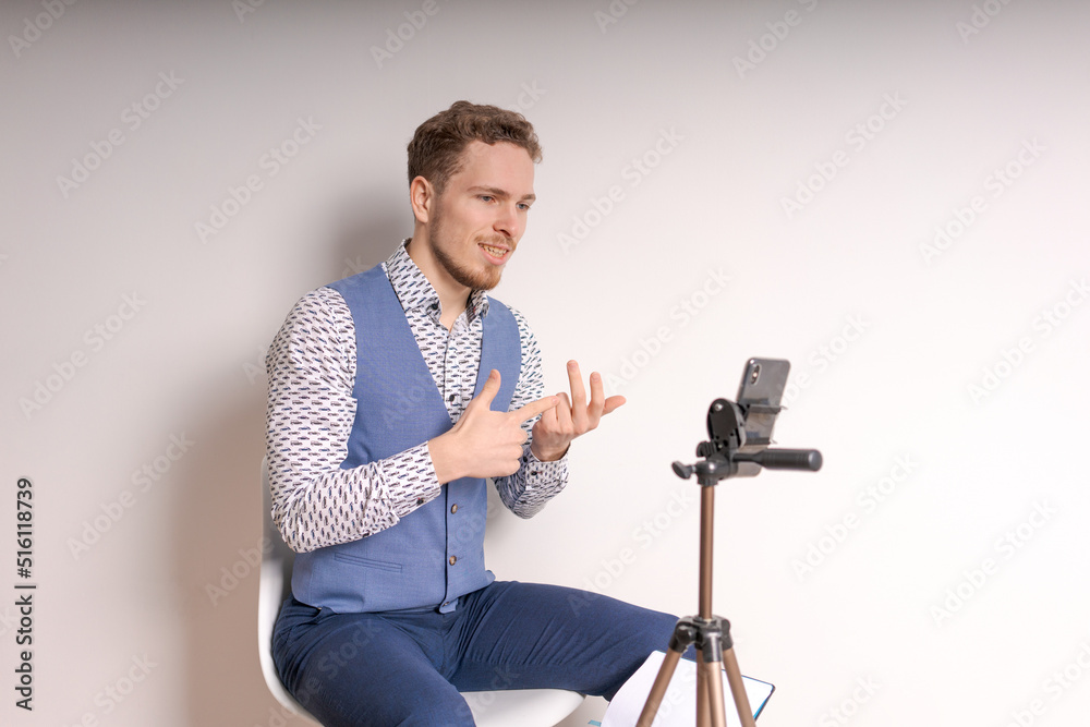 Young business man in shirt gesticulates while creating video in social networks online broadcast using a phone against a light wall, dressed in a shirt and a blue vest