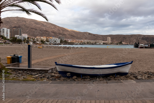 Panoramic view on sand beach of Playa de Los Cristianos on Tenerife  Canary Islands  Spain. A decorative wooden boat on the side of the touristic promenade street. Small barren hills and city view