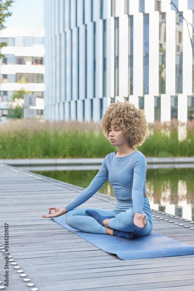 Calm fit woman sits in lotus pose on fitness mat practices yoga meditates for relaxation dressed in blue tracksuit poses near lake against cityscrapers. Feeling wellness and tranquility of mindfulness