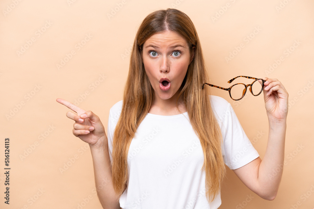 Young pretty blonde woman with glasses isolated on beige background surprised and pointing side Young pretty blonde woman with glasses isolated on beige background surprised and pointing side