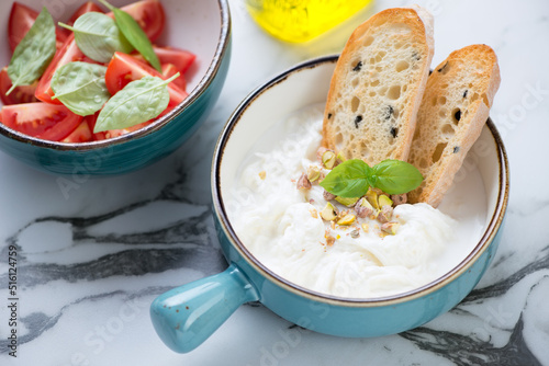 Turquoise bowl with stracciatella or stretched curd cheese, ciabatta and tomatoes, studio shot on a light-grey marble background, middle close-up