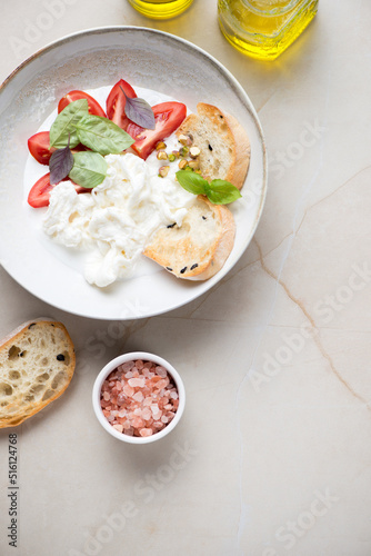 Stracciatella or cheese produced from italian buffalo milk, tomato wedges and ciabatta. Top view on a beige stone background, vertical shot with space
