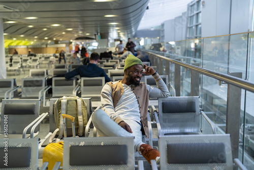 Smiling African American hipster man sitting in airport terminal or railway station, resting and looking at window. Happy passenger travelling with backpack waiting landing on board. 