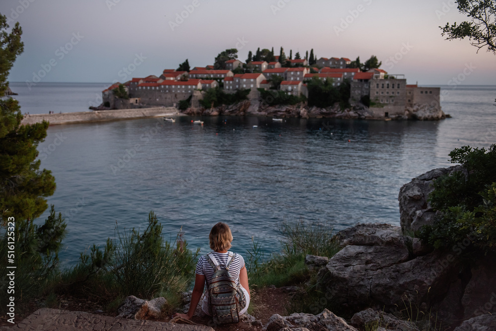 Adult pregnant woman in vest, white overalls stands by the sea. Traveler with a backpack looks at the island of Sveti Stefan at sunset in Montenegro. The concept of motherhood and pregnancy in travel