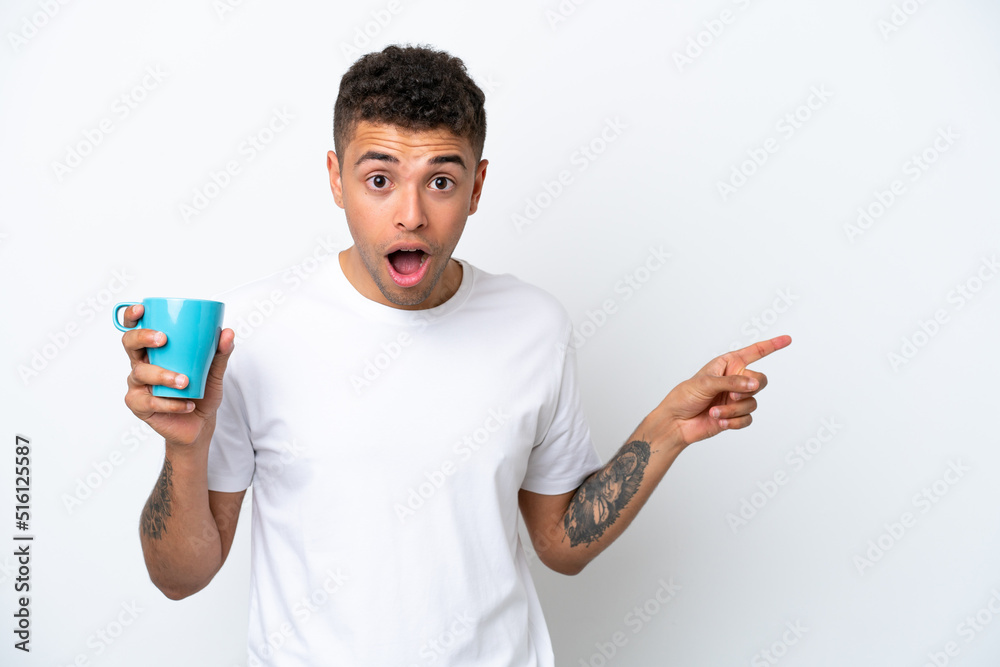 Young Brazilian man holding cup of coffee isolated on white background surprised and pointing finger to the side Young Brazilian man holding cup of coffee isolated on white background surprised and pointing finger to the side
