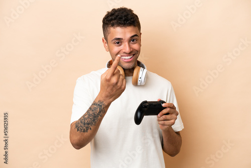 Young Brazilian man playing with a video game controller isolated on beige background doing coming gesture