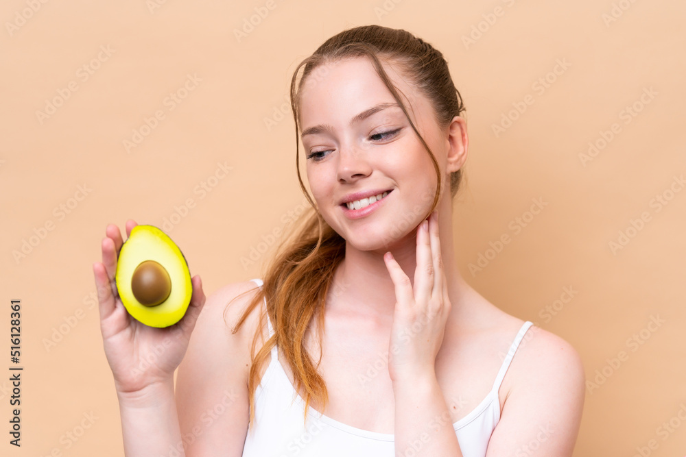 Young caucasian girl isolated on beige background holding an avocado while smiling. Close up portrait Young caucasian girl isolated on beige background holding an avocado while smiling. Close up portrait