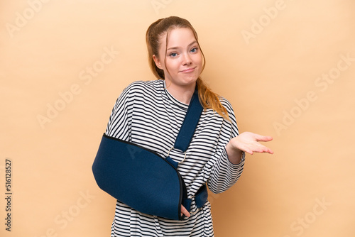 Young caucasian girl with broken arm and wearing a sling isolated on beige background making doubts gesture while lifting the shoulders photo