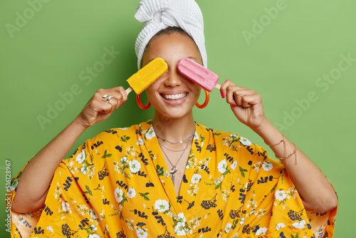 Horizontal shot of cheerful young woman covers eyes with delicious ice cream smiles broadly dressed in yellow robe white soft towel on head feels very happy isolated over bright green background