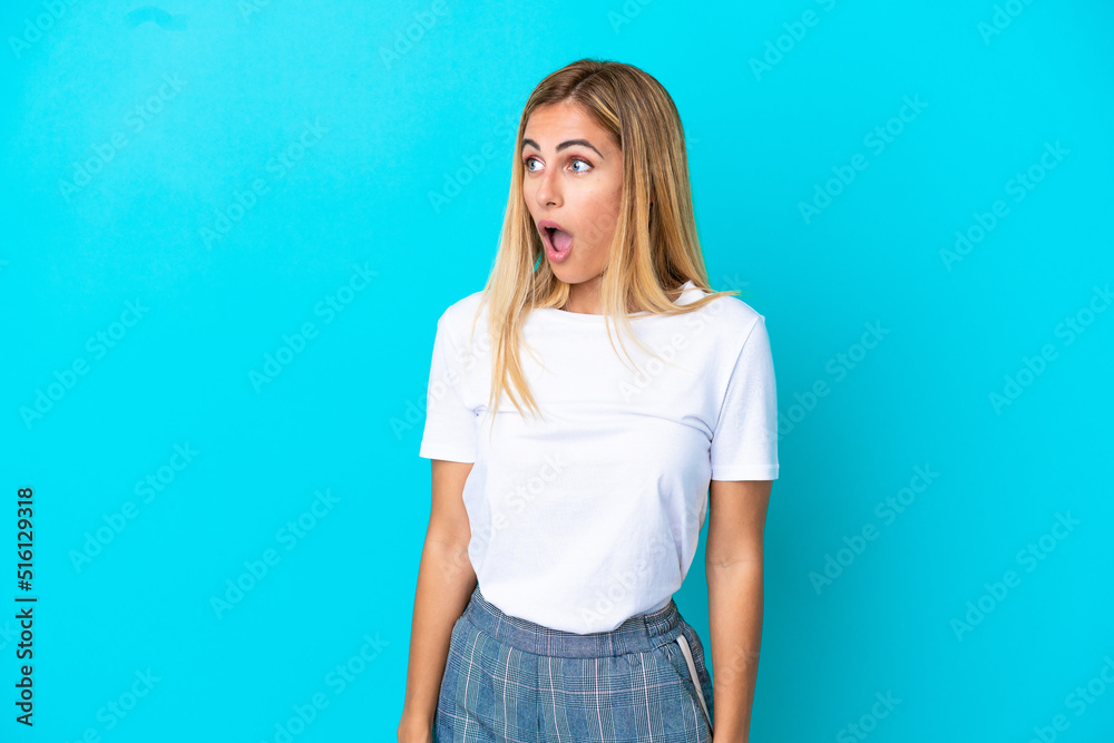 Blonde Uruguayan girl isolated on blue background doing surprise gesture while looking to the side Blonde Uruguayan girl isolated on blue background doing surprise gesture while looking to the side