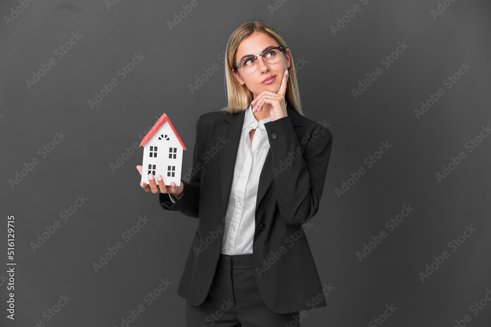 Blonde Uruguayan girl holding a house toy isolated on black background having doubts while looking up Blonde Uruguayan girl holding a house toy isolated on black background having doubts while looking up