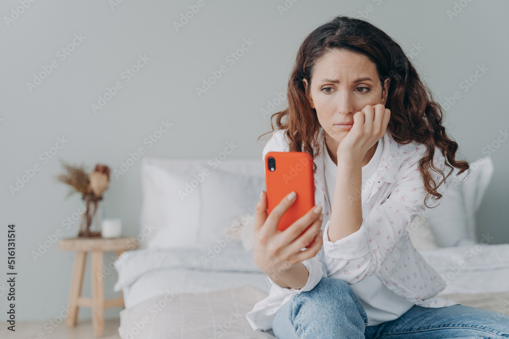 Stressed caucasian woman is doom scrolling. Girl reading morning news on smartphone in her bedroom. - obrazy, fototapety, plakaty 