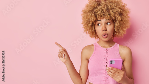 Impressed amazed woman with curly hair dressed in t shirt holds mobile phone points left on empty space shows awesome breathtaking advertisement isolated over pink background. Wow look at this