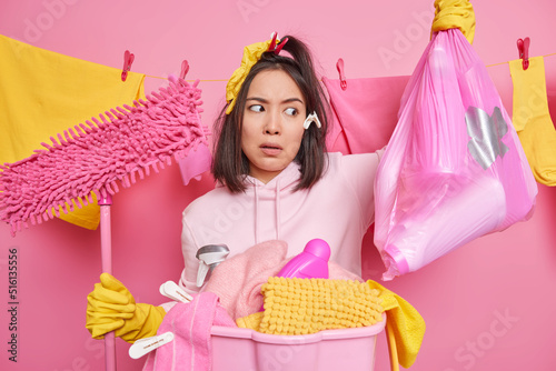 Shocked busy woman housekeeper looks at rubbish bag holds mop busy cleaning everything at home stands near basin full of laundry wears sweatshirt and rubber gloves isolated over pink background. photo