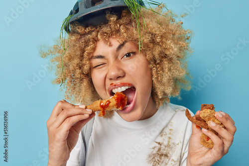 Curly haired millennial girl eats tasty fried nuggets with ketchup winks eye dressed in white t shirt and protective helmet prefers fast food containing much calories isolated over blue background
