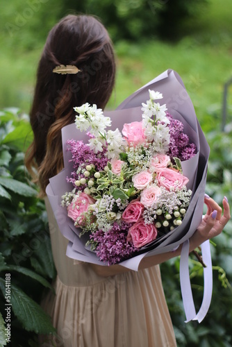 a delicate pink bouquet in the hands of a girl standing with her back in a light dress with a beautiful hairpin on her hair against a background of greenery