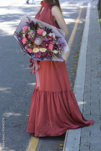 a girl in a long red dress with a large bouquet in her hands on the side of the road