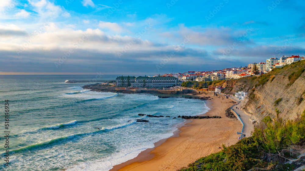 Aerial view - touristic coastline on summer while sunset. Beautiful seashore with skyline, waves and blue sky. Drone view over sandy beach  touristic city Ericeira , Portugal.