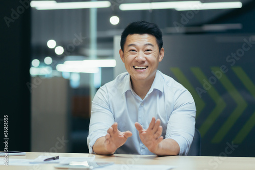 Successful and happy Asian businessman looking at camera, talking on video call, cheerfully gesticulating to interlocutor, online meeting, man working in office at work