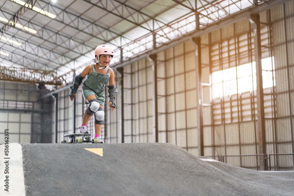 asian child or kid girl fun playing skateboard or surfskate and start on indoor pump track in skate park by extreme sports to wearing helmet elbow pads wrist and knee support for body safety on summer