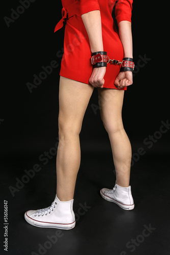 Young woman in short red dress stand of her back with leather handcuffs, on black background. Copy space for any text. Symbol of lack of freedom or struggle for rights, loving games, sensuality.