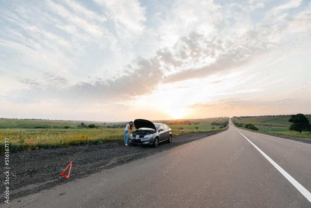 A young girl stands near a broken car in the middle of the highway during sunset and tries to repair it. Troubleshooting the problem. Waiting for help. Car service. Car breakdown on road..