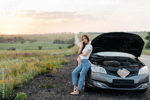 A frustrated young girl stands near a broken-down car in the middle of the highway during sunset. Breakdown and repair of the car. Waiting for help. Car service. Car breakdown on road. © Andrii