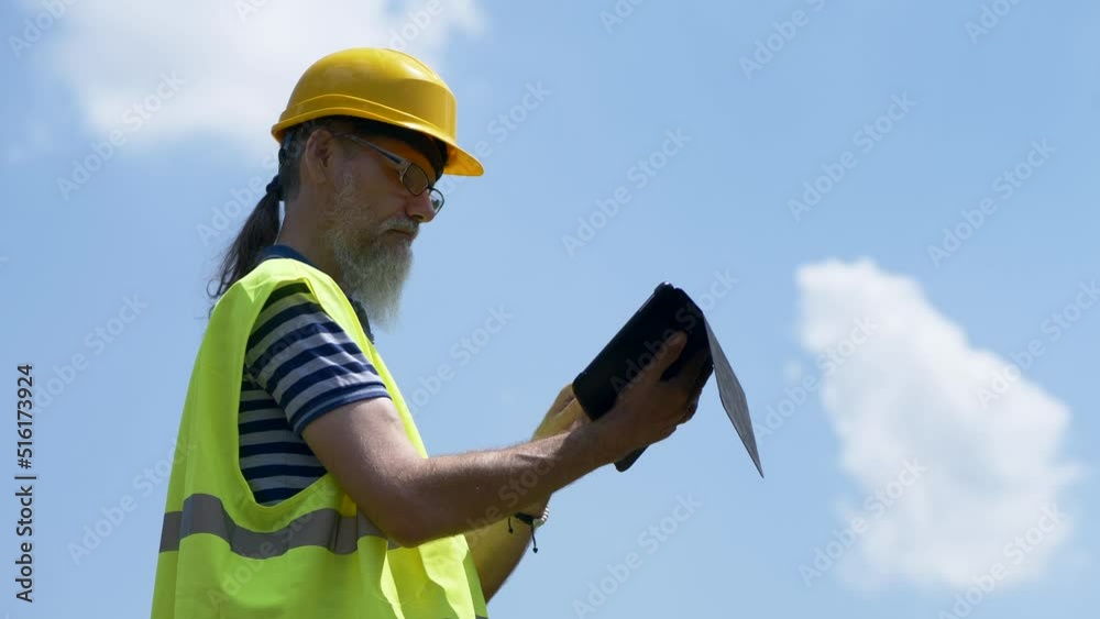 Port Worker With A Beard In A Yellow Helmet Stands With A Tablet PC. The Foreman Inspects. 4K Port Worker With A Beard In A Yellow Helmet Stands With A Tablet PC. The Foreman Inspects. 4K