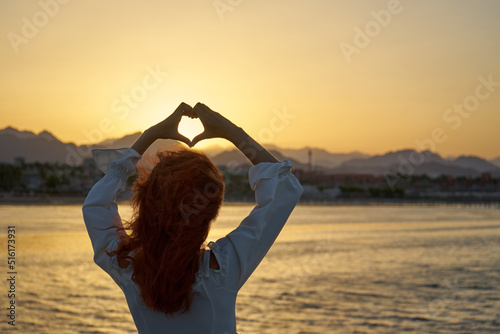Female making heart shape hand in a beautiful sunset nature setting at the beach.Love,life, romance. and compassion concept.Golden sunset with panoramic view of mountains.Travel and enjoy life