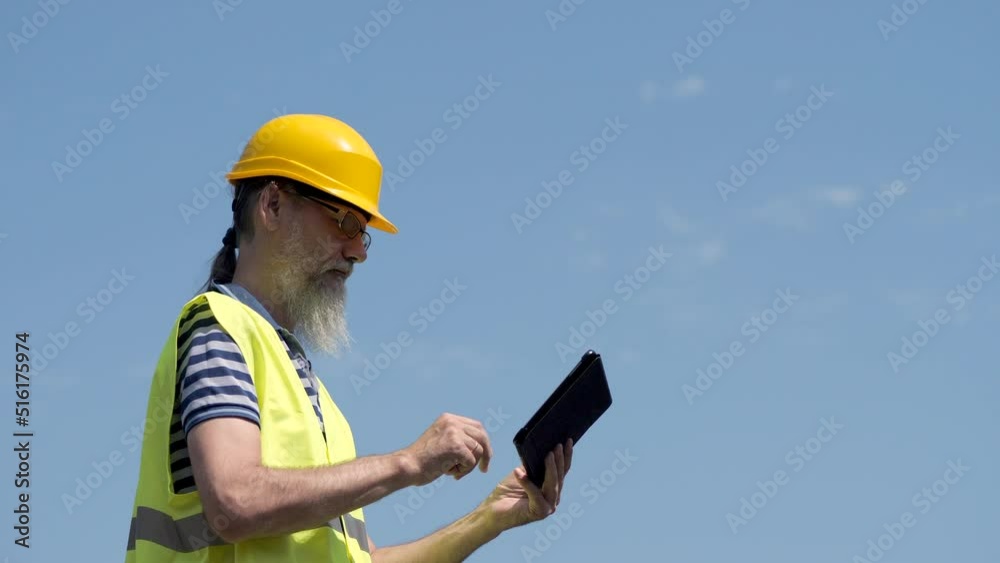 Port Worker With A Beard In A Yellow Helmet Stands With A Tablet PC. The Foreman Inspects. 4K Port Worker With A Beard In A Yellow Helmet Stands With A Tablet PC. The Foreman Inspects. 4K