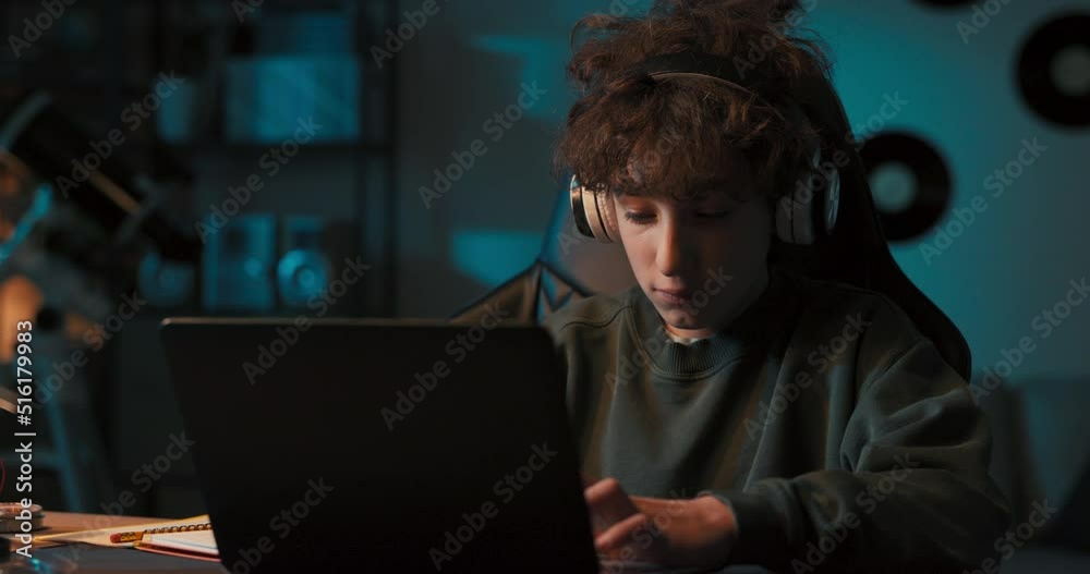 Smiling young college-aged boy has wireless headphones listens to music and works at computer, writes paper for school at desk at home. Smiling young college-aged boy has wireless headphones listens to music and works at computer, writes paper for school at desk at home.