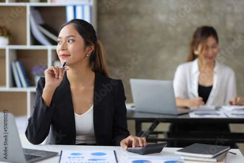 Happy Asian business woman on office desk working with laptop and looking away imagining success.